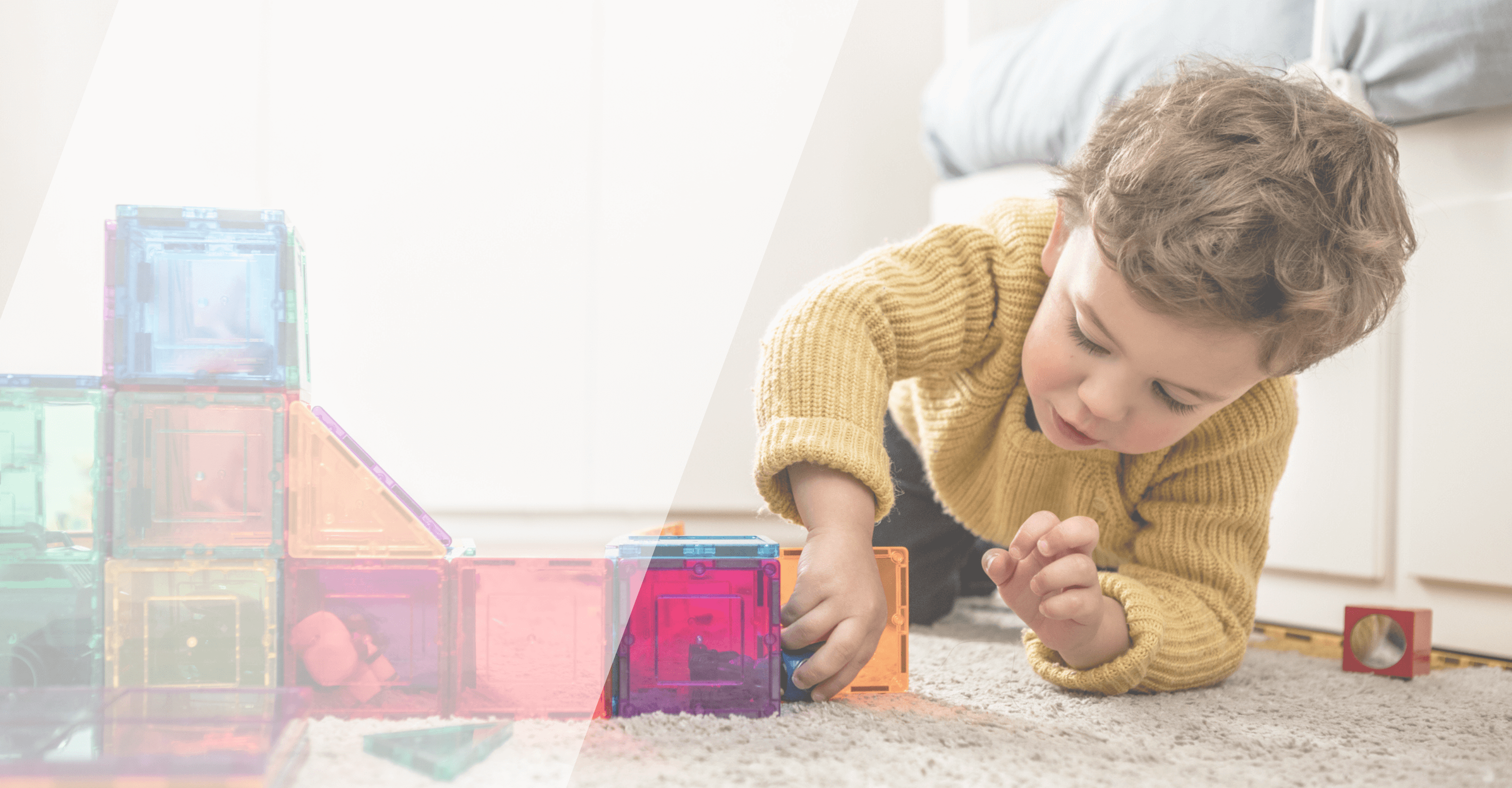 Jeune enfant jouant par terre avec des blocs colorés translucides, concentré sur sa construction. L'enfant porte un pull jaune moutarde et est dans une pièce lumineuse avec un sol en moquette claire.