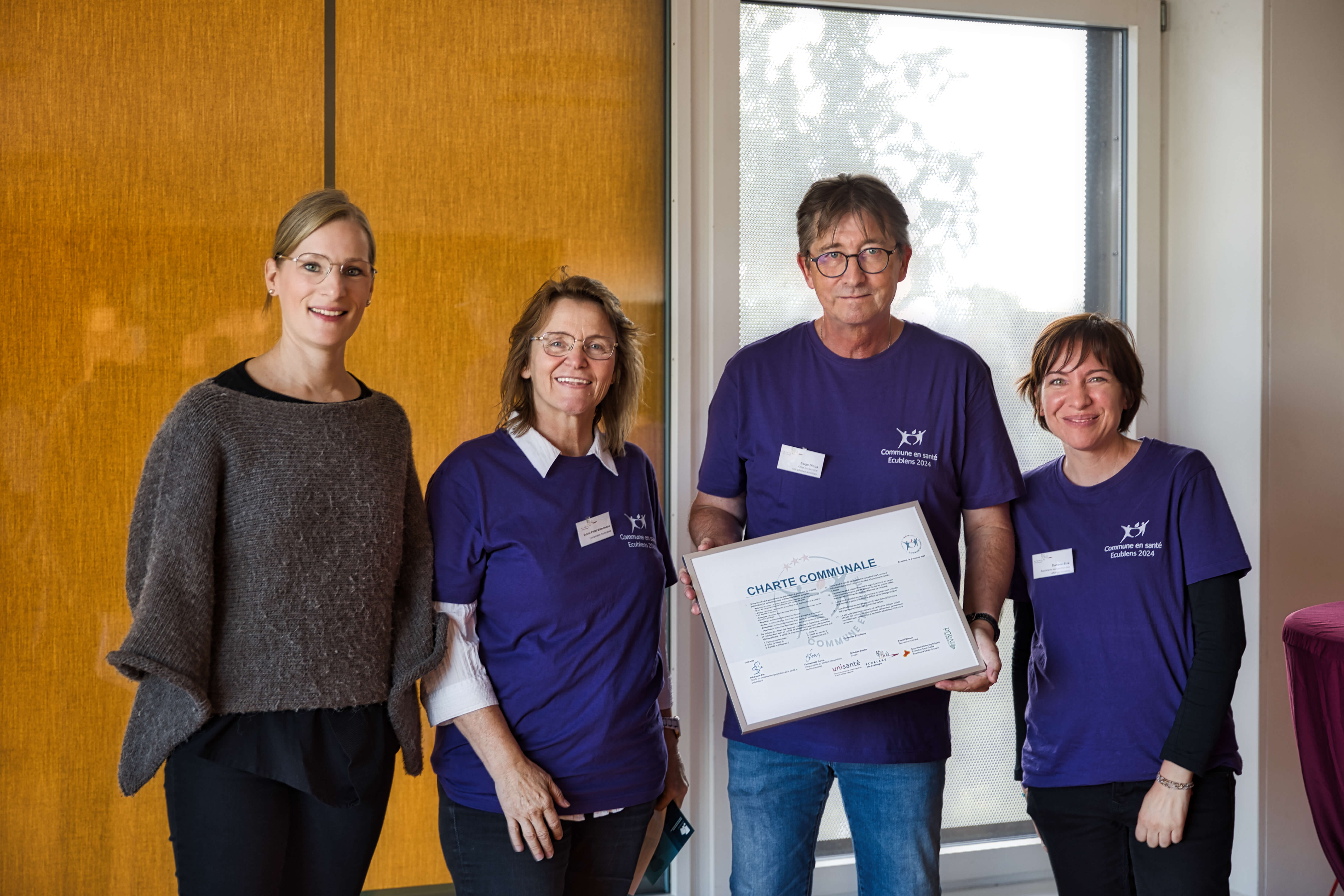 Quatre personnes souriantes posent ensemble, dont trois portant des t-shirts violets avec l'inscription 'Commune en santé Ecublens 2024'. L'une d'elles tient un cadre affichant une 'Charte communale'. Elles se trouvent dans une pièce avec un mur en bois à l'arrière-plan.