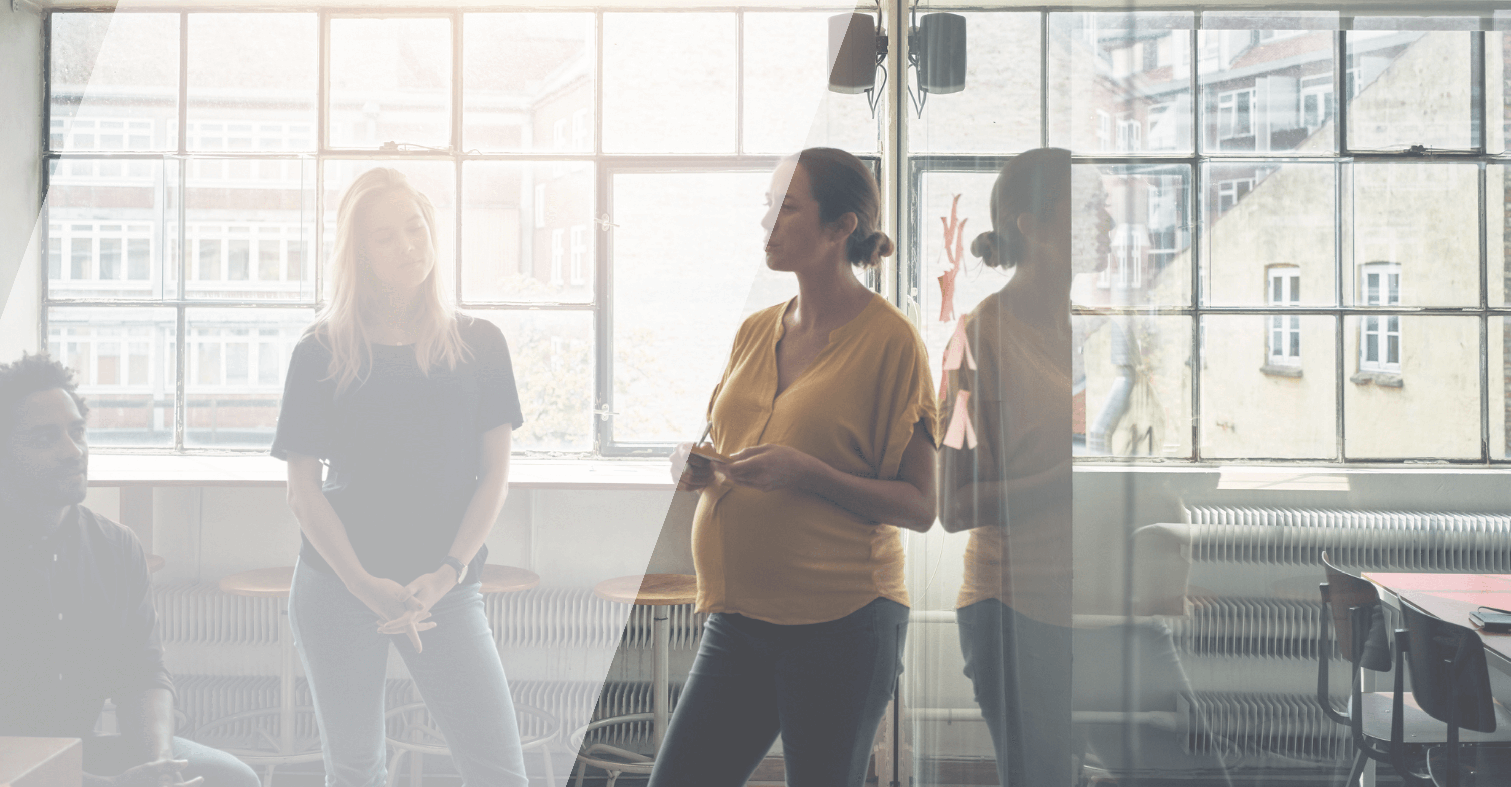 Scène de discussion dans un bureau lumineux, avec une femme enceinte debout en premier plan, portant un haut jaune, s'exprimant devant deux collègues assis. L'environnement est baigné de lumière naturelle provenant de grandes fenêtres, et des post-it sont visibles sur le mur en arrière-plan.