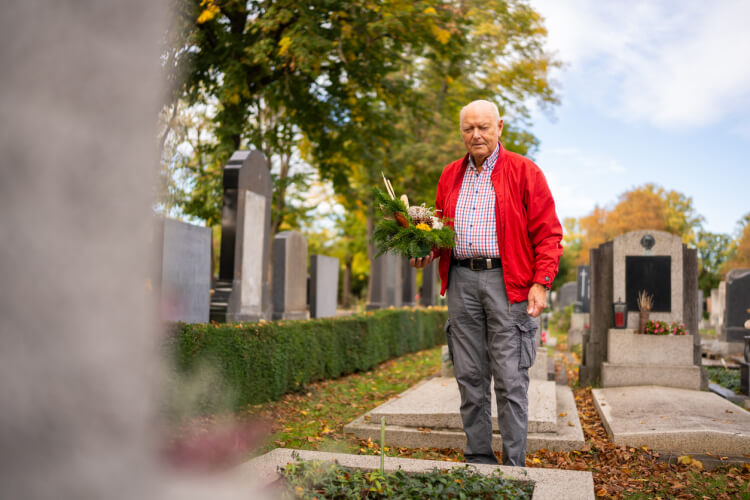 Homme âgé portant une veste rouge et tenant une composition florale dans un cimetière, debout devant une tombe. Les pierres tombales et les arbres aux feuilles automnales sont visibles à l'arrière-plan sous un ciel dégagé