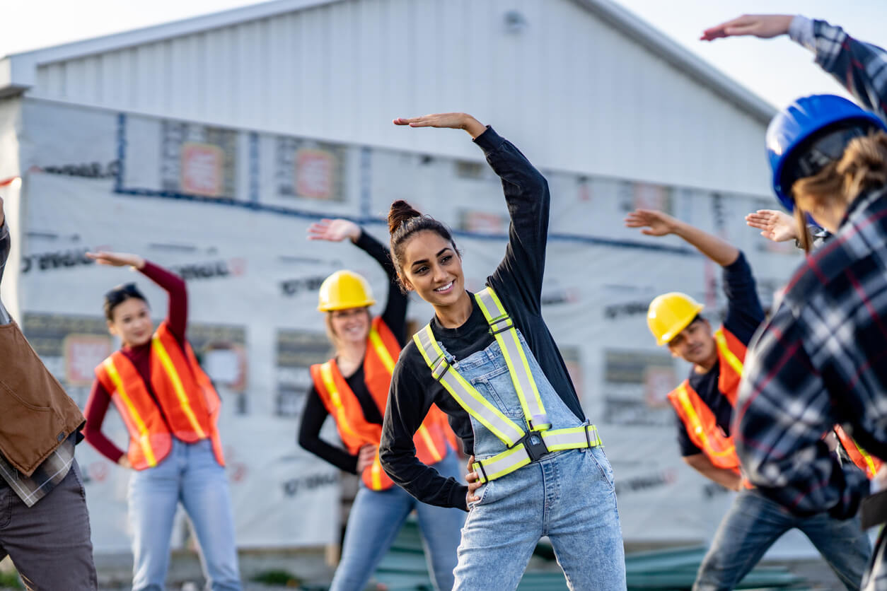 Groupe de travailleurs de la construction faisant des étirements ou des exercices d'échauffement sur un chantier. Ils portent des gilets de sécurité haute visibilité et des casques de protection. L'activité se déroule devant un bâtiment en construction