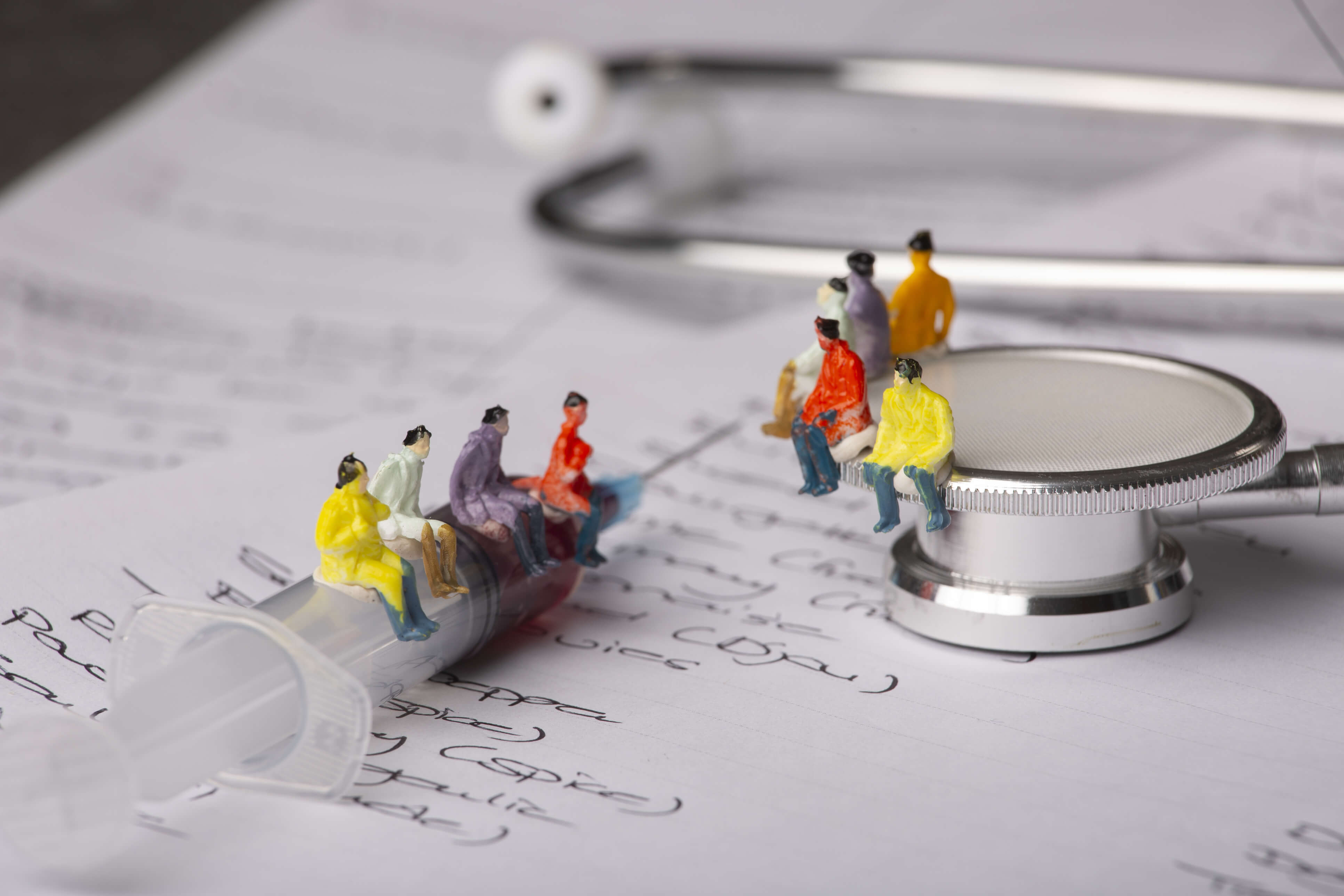 Photo de figurines miniatures assises sur une seringue et un stéthoscope, placées sur une feuille de papier avec des notes manuscrites, symbolisant l'impact des soins de santé sur les individus.